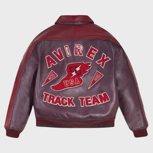 以下が含まれることがあります： 背面にAVIREX TRACK TEAMの赤い文字と白い縁取りの大きなグラフィックが施された、バーガンディ色のレザー製ボンバージャケット。デザインには、USA、1975、2035の文字が入った翼のある足のエンブレムが含まれています。ジャケットには襟、リブ編みの袖口、裾が付いています。