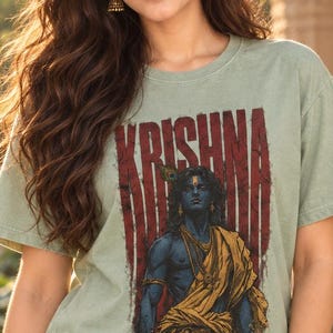 May include: A sage green short-sleeve t-shirt featuring a graphic print of the deity Krishna in blue skin with a yellow garment and peacock feather. The word KRISHNA is printed in large, distressed red capital letters above the figure.
