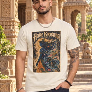 May include: A cream-coloured short-sleeve t-shirt featuring a rectangular graphic print on the chest. The print displays a blue-skinned figure in profile, surrounded by cosmic elements, lotus flowers, and the text "Hare Krishna" in a stylised font.