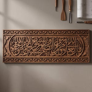 Puede incluir: Una placa de pared rectangular de madera con intrincada caligrafía árabe en relieve profundo, centrada dentro de un borde decorativo de motivos de enredaderas. La veta de madera marrón natural es visible en toda la superficie tallada.
