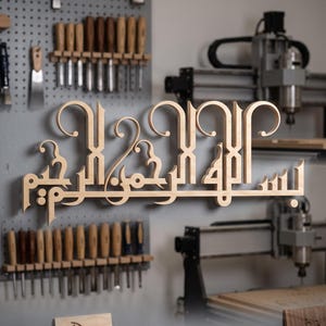 Puede incluir: Un letrero de pared de madera de color claro con una intrincada caligrafía árabe. La escritura está cortada con láser con bordes lisos y un acabado de grano de madera natural. El diseño muestra la frase Bismillah ir-Rahman ir-Rahim.
