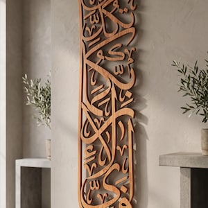 May include: A vertical wooden wall art piece featuring intricate, laser-cut Arabic calligraphy. The natural brown wood grain is visible throughout the detailed, flowing script. The piece is mounted flat against a neutral-coloured wall surface.