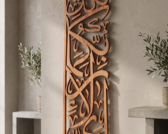 May include: A vertical wooden wall art piece featuring intricate, laser-cut Arabic calligraphy. The natural brown wood grain is visible throughout the detailed, flowing script. The piece is mounted flat against a neutral-colored wall surface.