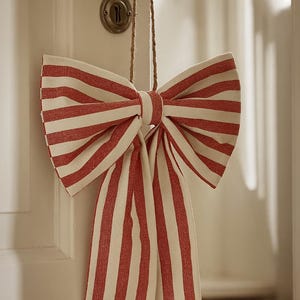 May include: A decorative fabric bow featuring a pattern of vertical red and off-white stripes. The bow is constructed with two large loops and two long, hanging tails, and it is suspended by a thin, natural-colored twine loop for hanging.