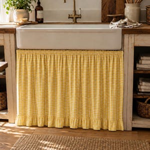 May include: A yellow and white gingham fabric sink skirt featuring a gathered top and a ruffled bottom hem. The curtain is installed beneath a white farmhouse-style sink, covering the open cabinet space with a pleated, checkered textile design.