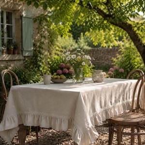 Op de afbeelding: Een rechthoekig beige linnen tafelkleed met een geplooide rand. Het tafelkleed ligt over een tafel die is gedekt met een doorzichtige glazen vaas met witte en gele wilde bloemen, een keramische mok, een schaal met fruit en een kleine lantaarn.