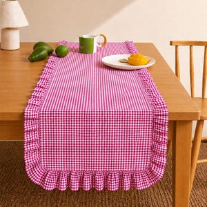 May include: A rectangular table runner featuring a vibrant pink and white gingham check pattern. The runner is finished with a gathered ruffle trim along all edges, creating a decorative border that drapes over the ends of a wooden dining table.