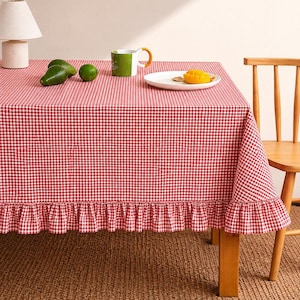 May include: A red and white gingham tablecloth with a ruffled edge covers a wooden table. A green mug, a white plate with a mango slice, and fresh avocados and limes are on the table. A small lamp is on the table.