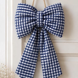 May include: A decorative bow made of navy blue and white gingham fabric. The bow features two large loops and two long, hanging tails. A thin, light brown jute twine loop is attached to the top center of the bow for hanging purposes.