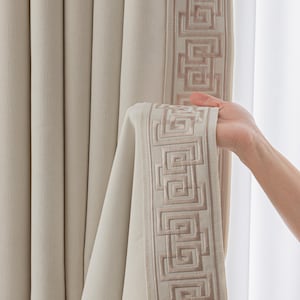 Puede incluir: Paneles de cortina beige con un borde bordado geométrico en un tono más claro. Las cortinas están hechas de una tela gruesa y texturizada y cuelgan frente a una ventana.