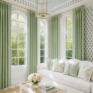 May include: A white sofa with sage green and white patterned throw pillows, sage green curtains with a Greek key trim, a white coffee table with a glass vase of flowers and stacked books, and a gold lantern pendant light fixture.