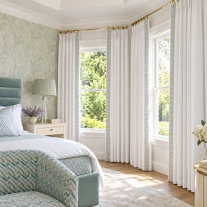 May include: Floor-to-ceiling white curtains with a decorative light blue patterned border hanging on a brass rod. The curtains frame two windows in a bedroom featuring a light blue upholstered bed, a bedside table, and a lamp.