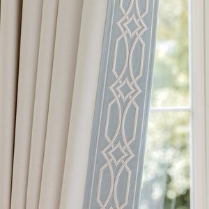 May include: A pleated curtain panel in a solid off-white color featuring a vertical border trim. The trim is light blue with a white embroidered geometric pattern consisting of repeating interlocking shapes and diamond outlines along the edge.