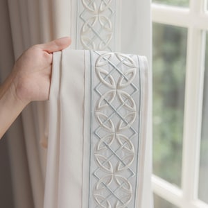 May include: A cream-colored curtain panel featuring a vertical border with an embroidered geometric pattern. The design consists of repeating circular and diamond shapes stitched in light blue and off-white thread on a textured fabric base.