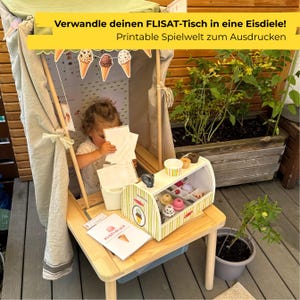 Könnte beinhalten: Ein hölzerner Spieltisch mit Stoffüberdachung und einem Eisdielen-Spielset mit verschiedenen Eiskugeln und Bechern. Das Set enthält bedruckte Papierschilder mit Eistüten-Illustrationen und dem Text Verwandle deinen FLISAT-Tisch in eine Eisdiele!