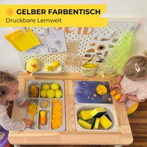 Könnte beinhalten: Ein Holztisch mit gelben und grauen Fächern, gefüllt mit Spielzeug und Bastelmaterialien. Der Tisch hat eine weiße Lochwand mit Zeichnungen und ein gelbes Banner mit der Aufschrift "GELBER FARBENTISCH Druckbare Lernwelt".
