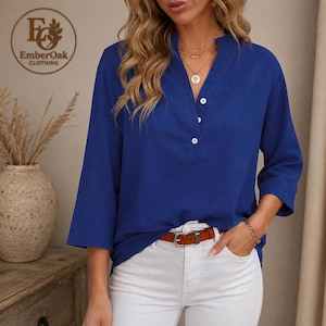 Op de afbeelding: Een kobaltblauwe blouse met een opstaande kraag en drie witte knopen. De top met driekwartmouwen wordt gecombineerd met een witte broek en een bruine riem. Het EmberOak Clothing-logo is linksboven zichtbaar.