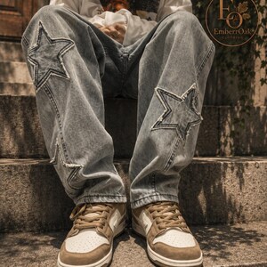 May include: Light wash denim jeans featuring star-shaped patches on the knees. The jeans are worn with brown and white trainers. The image shows a person sitting on concrete steps, highlighting the jeans' design and fit. The jeans have a relaxed, casual style.