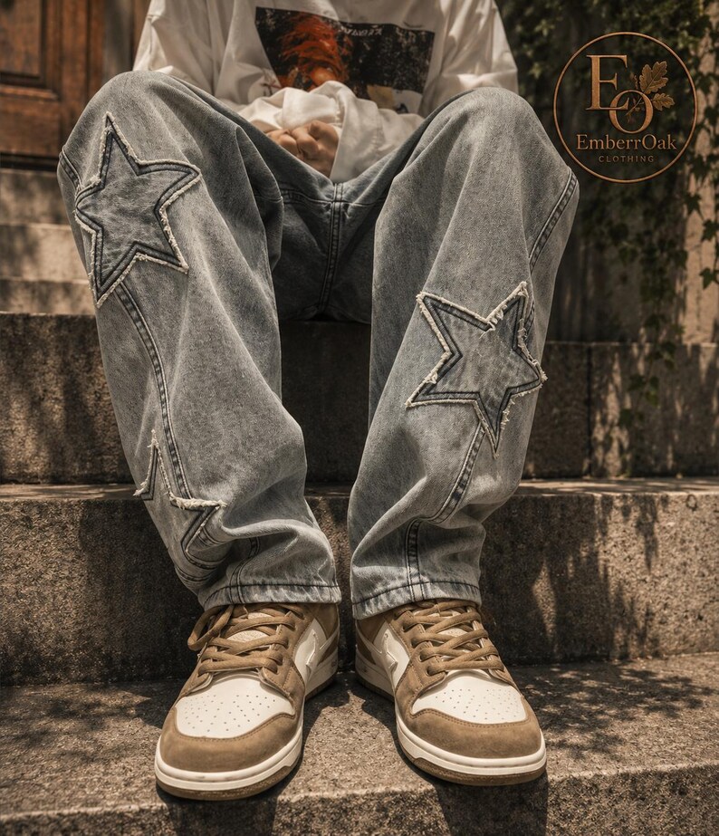 May include: Light wash denim jeans featuring star-shaped patches on the knees. The jeans are worn with brown and white trainers. The image shows a person sitting on concrete steps, highlighting the jeans' design and fit. The jeans have a relaxed, casual style.