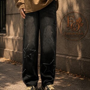 May include: Black denim jeans featuring large star cutouts and distressed edges. The jeans are worn with tan and black trainers. A brown long-sleeve top is also visible. The EmberrOak Clothing logo is in the background.