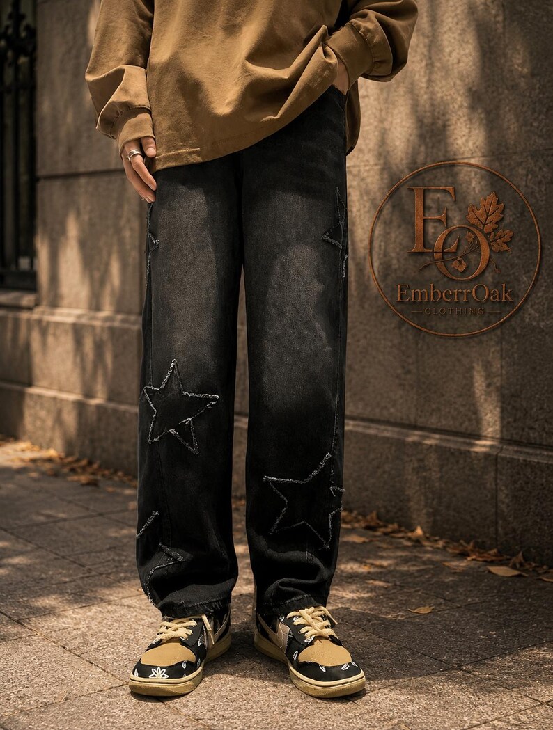 May include: Black denim jeans featuring large star cutouts and distressed edges. The jeans are worn with tan and black trainers. A brown long-sleeve top is also visible. The EmberrOak Clothing logo is in the background.