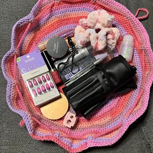 May include: A round, multi-colored crochet drawstring bag in shades of pink, orange, and purple, containing various beauty items including a Kiss magnetic nail set, a black makeup brush roll, a BareMinerals compact, a hair scrunchie, and tools.