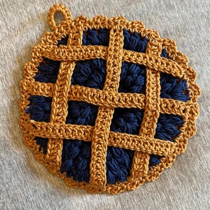 Peut inclure: Un dessous-de-plat rond au crochet en forme de tarte avec un motif de croûte en treillis. La base est faite de fil bleu marine foncé, tandis que le dessus en treillis et la bordure festonnée sont réalisés en fil brun doré. Une boucle est fixée.