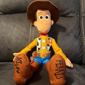 Puede incluir: Mu&ntilde;eco de peluche de Woody de la franquicia Toy Story. El mu&ntilde;eco tiene un sombrero de vaquero marr&oacute;n, una camisa a cuadros amarilla, un chaleco con estampado de vaca, pantalones azules y botas marrones. La palabra "ANDY" est&aacute; escrita en una bota y "FERRIS" en la otra.