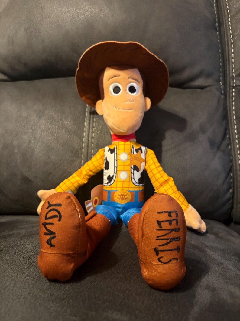 Puede incluir: Mu&ntilde;eco de peluche de Woody de la franquicia Toy Story. El mu&ntilde;eco tiene un sombrero de vaquero marr&oacute;n, una camisa a cuadros amarilla, un chaleco con estampado de vaca, pantalones azules y botas marrones. La palabra "ANDY" est&aacute; escrita en una bota y "FERRIS" en la otra.