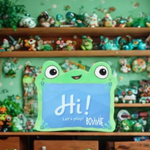 Può includere: Un cartello verde a forma di rana con la scritta "Hi!" in bianco e "Let's play!" in caratteri più piccoli. Il cartello ha un centro blu ed è circondato da una collezione di statuette di rane sugli scaffali.