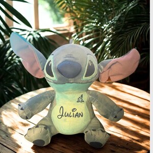 May include: A soft toy Stitch doll in blue, grey, and green, with large ears and the name "Julian" embroidered on its chest. The doll is on a wooden surface, with a leafy green background.