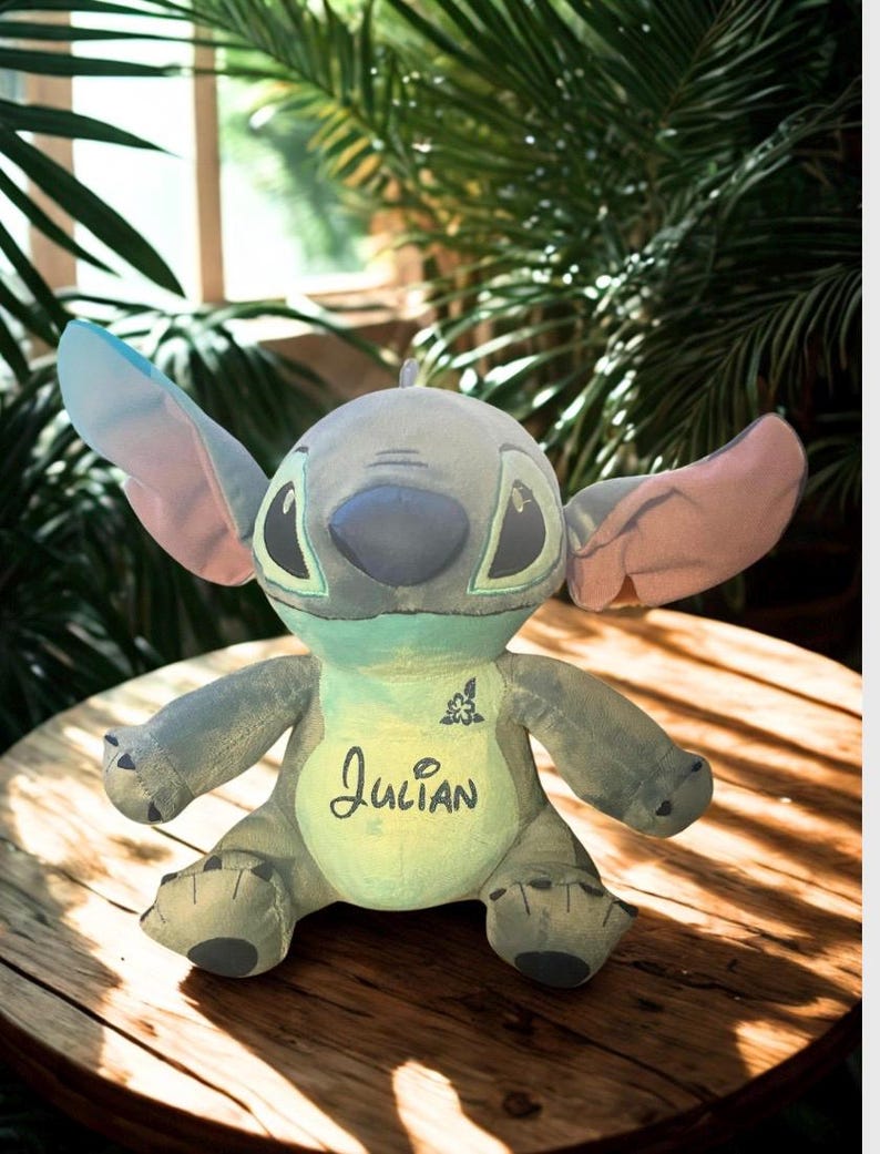May include: A soft toy Stitch doll in blue, grey, and green, with large ears and the name "Julian" embroidered on its chest. The doll is on a wooden surface, with a leafy green background.