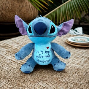May include: A blue Stitch plush toy with large ears and a dark blue nose. The toy has a heart and text in Portuguese on its chest. It is sitting on a woven surface.