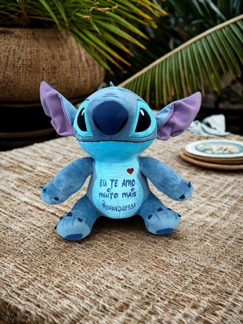 May include: A blue Stitch plush toy with large ears and a dark blue nose. The toy has a heart and text in Portuguese on its chest. It is sitting on a woven surface.