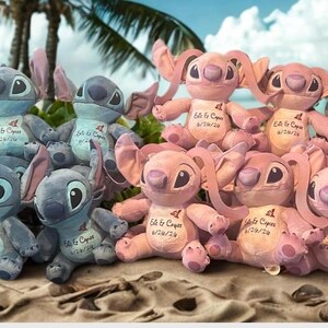 May include: A display of blue and pink Stitch plush toys, each with large ears and eyes, arranged on a sandy beach. The toys have tags and are set against a tropical backdrop of palm trees and the sea.
