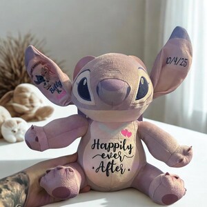 May include: A lavender Stitch plush toy with large ears and black eyes. The doll features the text "Happily ever After" on its chest, with two pink hearts. The left ear has a graphic of Stitch and a name, and the right ear has the date "10/4/25".