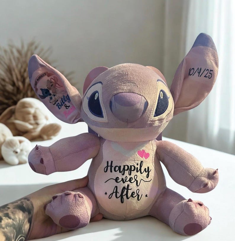 May include: A lavender Stitch plush toy with large ears and black eyes. The doll features the text "Happily ever After" on its chest, with two pink hearts. The left ear has a graphic of Stitch and a name, and the right ear has the date "10/4/25".