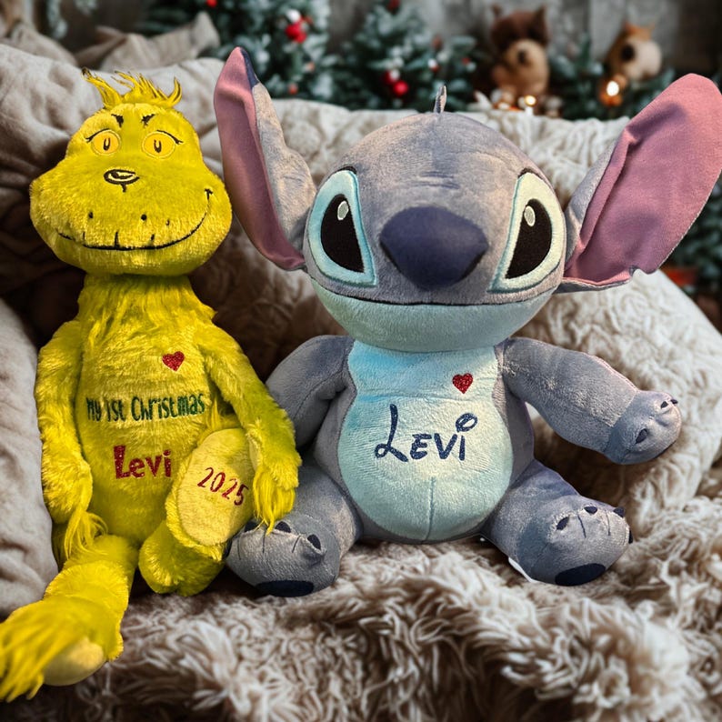 May include: Two plush toys: a yellow Grinch-like character and a blue Stitch-like character. The Grinch toy has the text "My 1st Christmas Levi 2025". The Stitch toy has the name "Levi" on its chest. Both toys are on a textured surface.