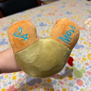 Puede incluir: Una almohada de felpa en forma de U, con un cuerpo verde claro y secciones anaranjadas en forma de oreja. El texto "DA" est&aacute; bordado en azul en una oreja y "Moy" en la otra. La almohada es suave y de material afelpado.