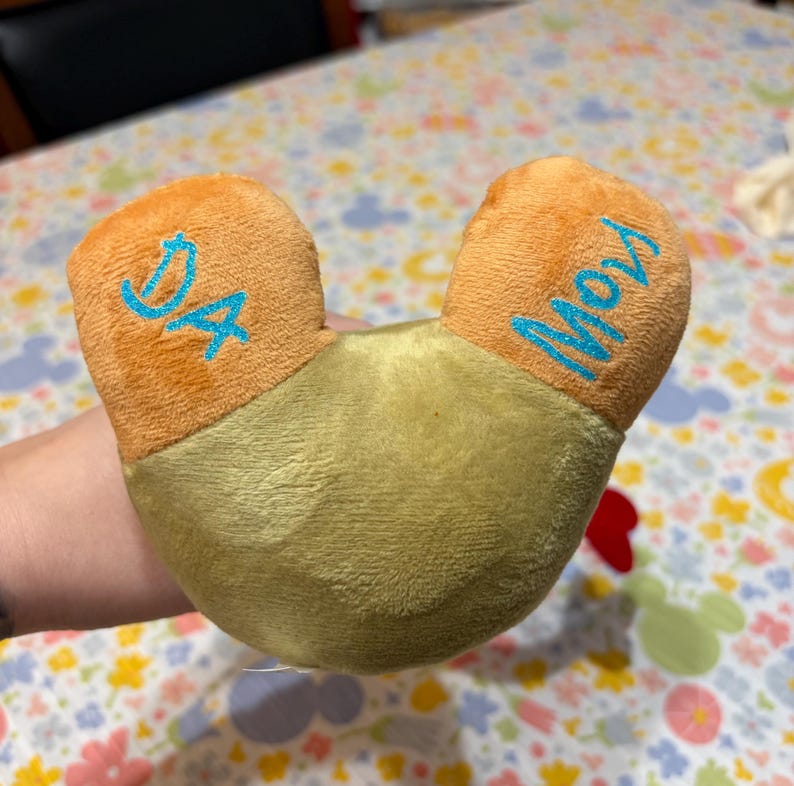 Puede incluir: Una almohada de felpa en forma de U, con un cuerpo verde claro y secciones anaranjadas en forma de oreja. El texto "DA" est&aacute; bordado en azul en una oreja y "Moy" en la otra. La almohada es suave y de material afelpado.