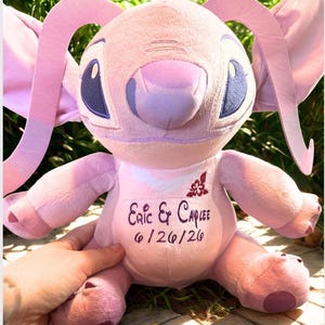 May include: A pink plush toy with large ears, blue eyes, and a lavender nose. The toy has the names "Eric & Caylee" and the date "6/26/20" embroidered on its chest. The toy is held by a hand.