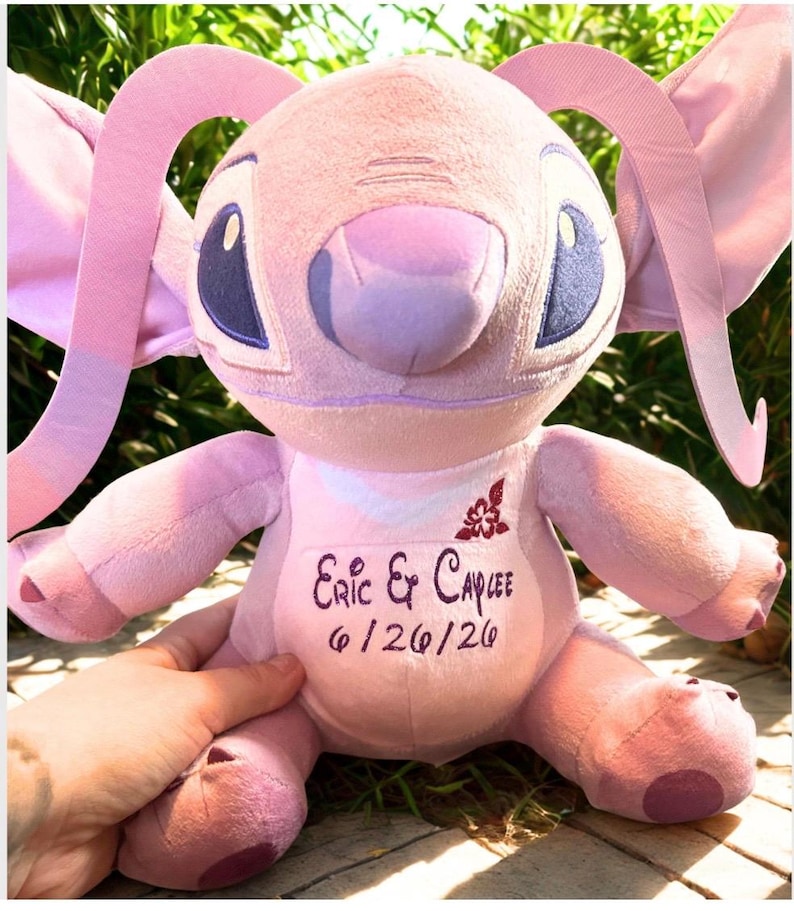 May include: A pink plush toy with large ears, blue eyes, and a lavender nose. The toy has the names "Eric & Caylee" and the date "6/26/20" embroidered on its chest. The toy is held by a hand.