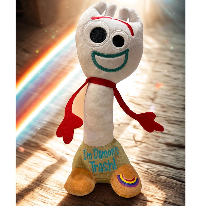 Puede incluir: Un peluche de Forky de Toy Story 4. El personaje blanco en forma de tenedor tiene grandes ojos negros, una sonrisa verde y una bufanda roja. La base beige tiene el texto "I'm Damon's Trash!" y un dise&ntilde;o de arco&iacute;ris.
