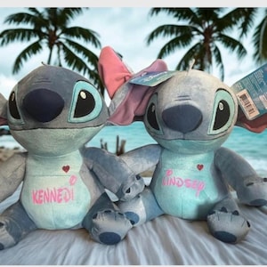 May include: Two blue and grey Stitch plush toys, one with a pink bow, are displayed against a beach scene. The dolls have large black eyes and the names "Kennedy" and "Lindsey" are embroidered on their chests. One doll has a tag attached.