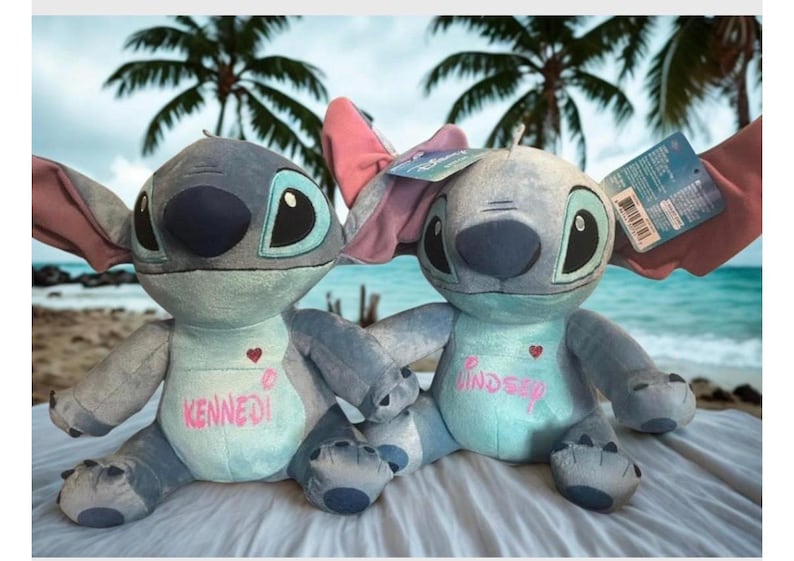 May include: Two blue and grey Stitch plush toys, one with a pink bow, are displayed against a beach scene. The dolls have large black eyes and the names "Kennedy" and "Lindsey" are embroidered on their chests. One doll has a tag attached.