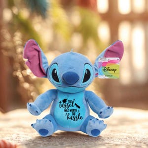 May include: A blue Stitch plush toy with large ears and a graduation message on its chest. The text reads "The tassel was worth the hassle." The toy has a Disney tag attached.