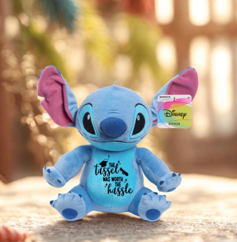 May include: A blue Stitch plush toy with large ears and a graduation message on its chest. The text reads "The tassel was worth the hassle." The toy has a Disney tag attached.