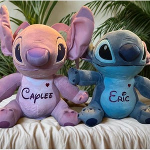 May include: Two plush toys of cartoon characters, one pink and one blue. The pink toy has the name "Caplee" embroidered on its chest, and the blue toy has the name "Eric". Both toys have large ears and are sitting on a white surface.