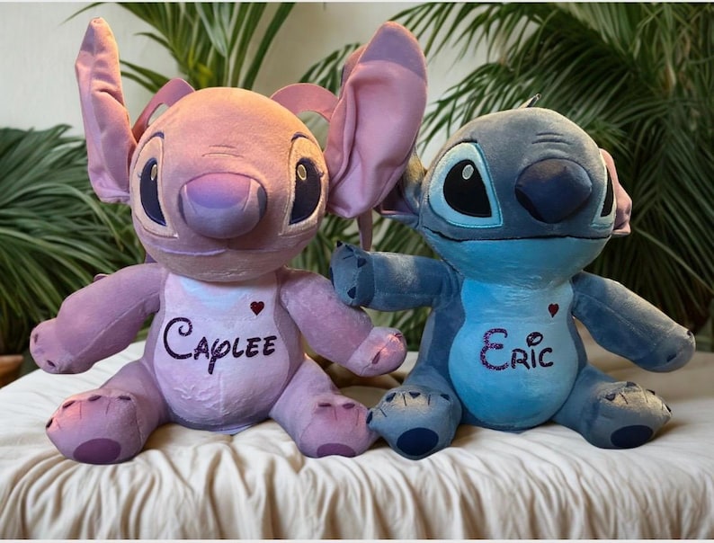 May include: Two plush toys of cartoon characters, one pink and one blue. The pink toy has the name "Caplee" embroidered on its chest, and the blue toy has the name "Eric". Both toys have large ears and are sitting on a white surface.