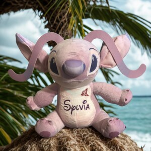 May include: A lavender-coloured plush toy with large ears and a smiling face. The word "Sylvia" is embroidered on the chest. The toy is sitting on a bed of straw, with a tropical background of palm leaves and ocean.